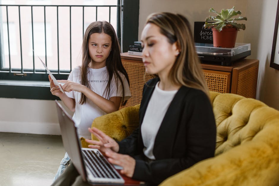 A woman in a black blazer uses a laptop on a couch while a young girl stands near holding paper.
