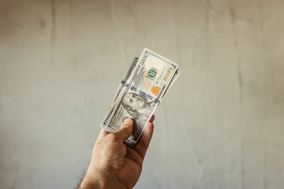 A hand holding a stack of US dollar banknotes against a neutral backdrop, symbolizing wealth.