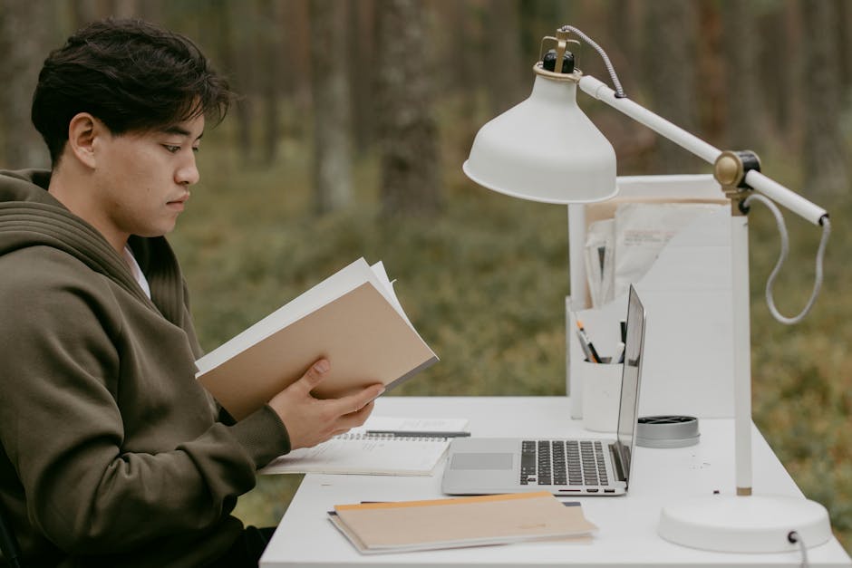 Asian man reading at an outdoor workspace with laptop, books, and lamp on desk.