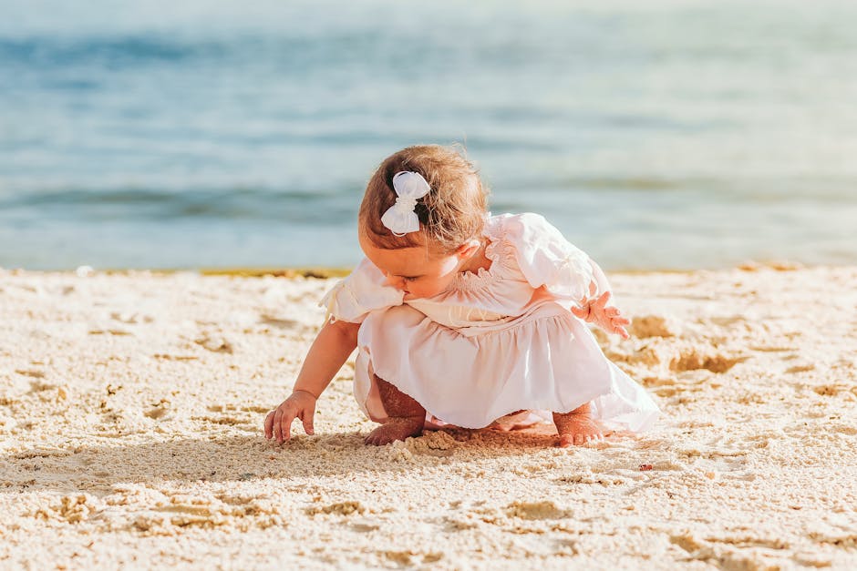 Full body adorable little girl in white summer dress sitting on haunches and playing with warm sand on sunny seacoast