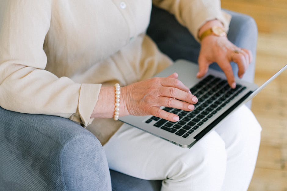 From above of crop anonymous elderly female typing information for report on netbook while resting in armchair in living room