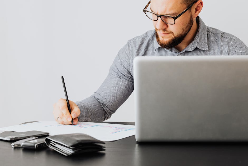 A focused man analyzing financial papers with a laptop in an office setting.