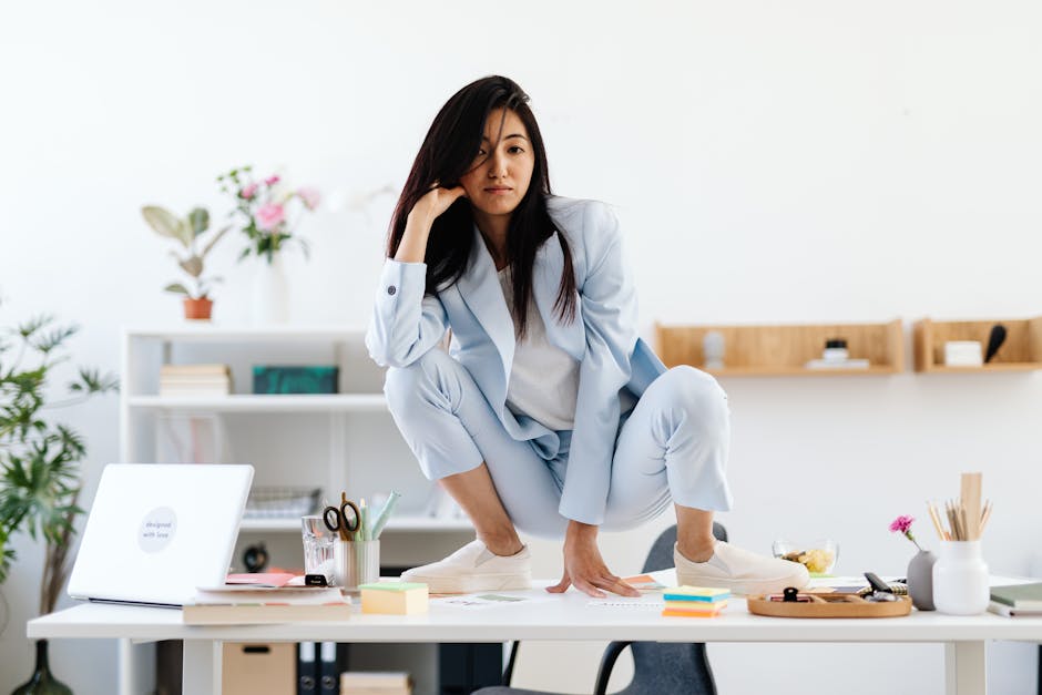 Asian woman in a blue suit poses confidently on an office table, surrounded by a modern workspace.