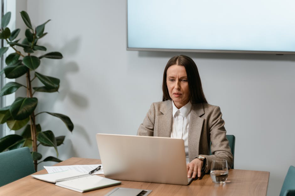 A woman in a business suit using a laptop at an office desk with documents and an indoor plant.