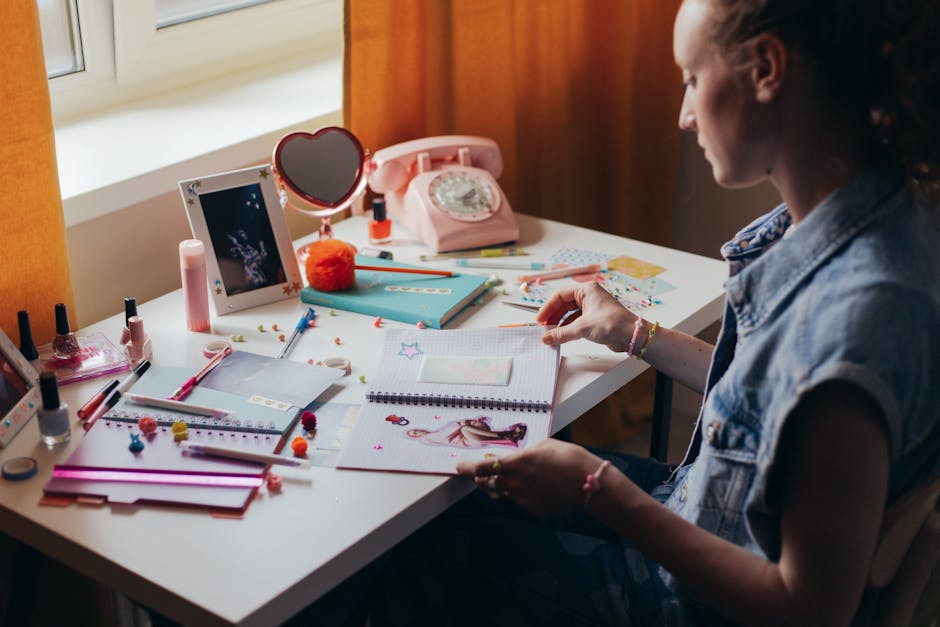 A woman sits at a cluttered desk working on a creative journal, surrounded by stationery and decorative items.