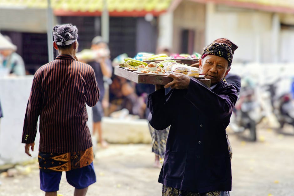 An Indonesian street vendor carries a tray of local delicacies at an outdoor market, showcasing cultural attire.