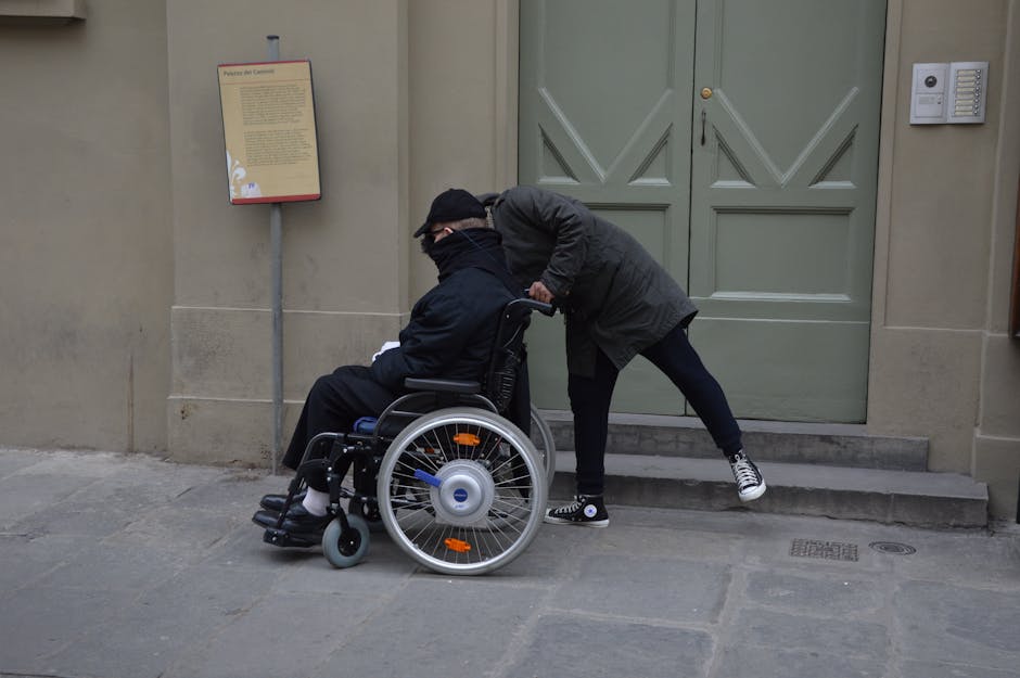 A caregiver assists a person in a wheelchair near a large door in a city street scene.