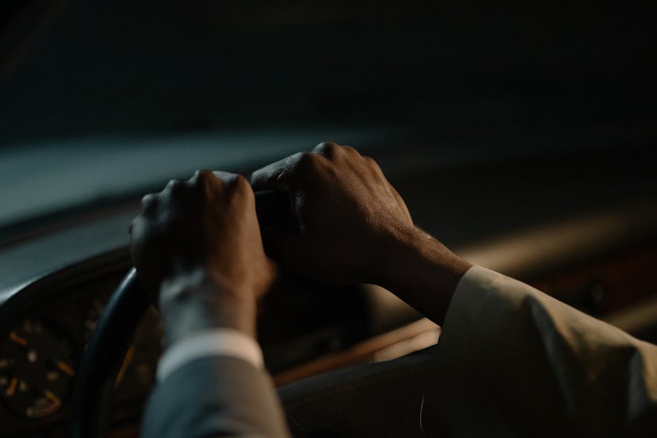 A driver's hands grasp the steering wheel in a dimly-lit vehicle interior, conveying focus and control.