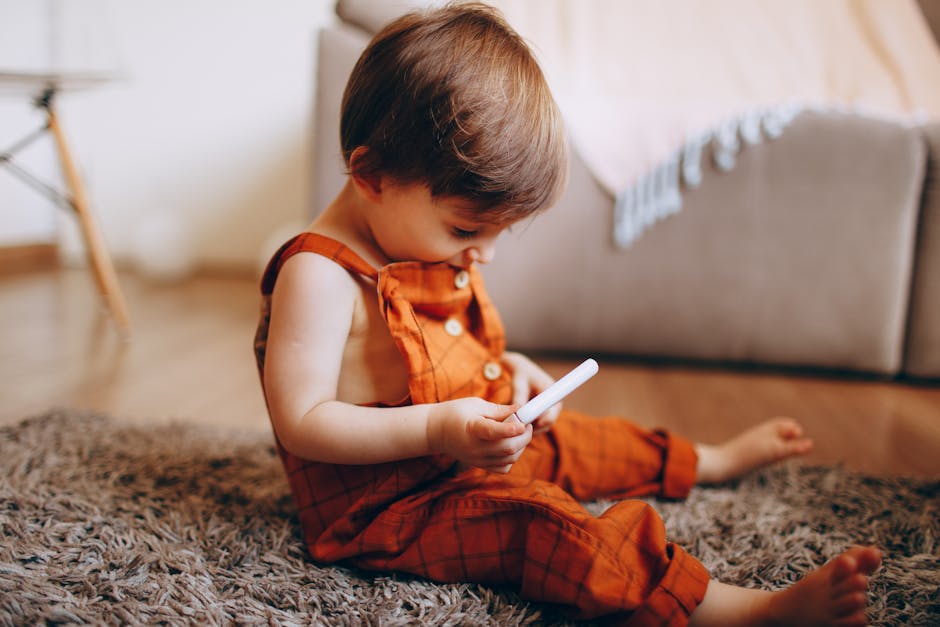 A cute toddler in orange overalls explores a toy while sitting on a rug indoors.