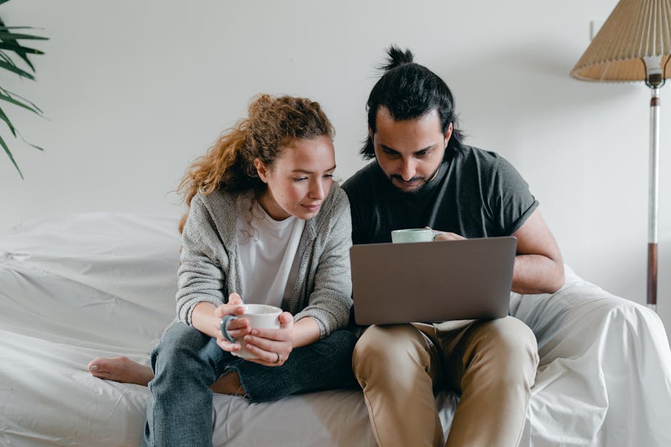 Positive young couple in casual clothes sitting on sofa with white cover and surfing netbook