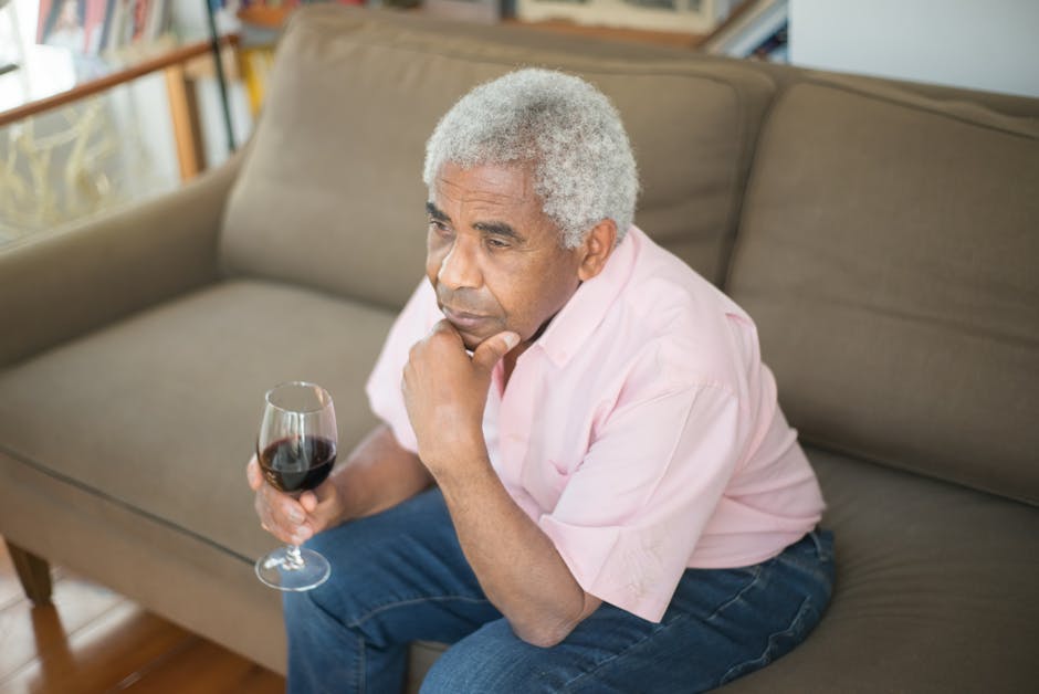 Contemplative elderly man sitting on a sofa holding a glass of wine indoors.