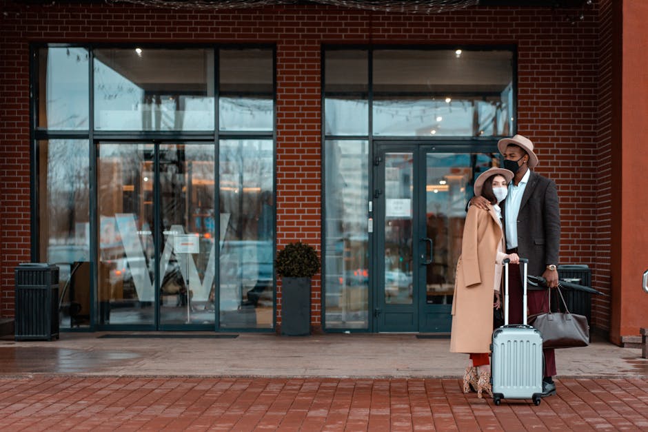 A fashionable couple in masks standing with luggage at a modern hotel entrance.