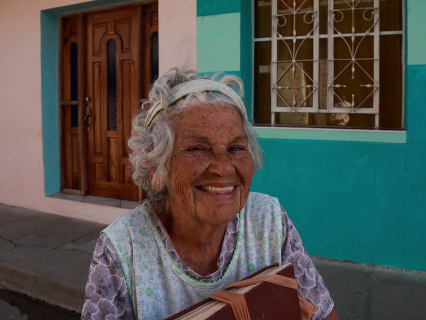 Joyful elderly woman with grey hair smiling and holding a book outside a colorful home.