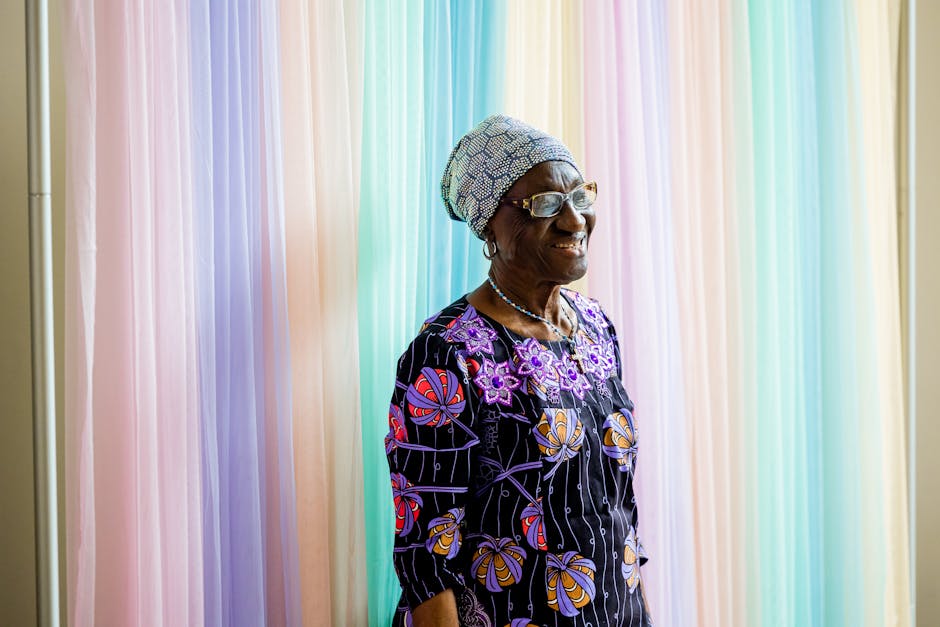 Elderly woman with glasses and headwrap, standing against a pastel background.