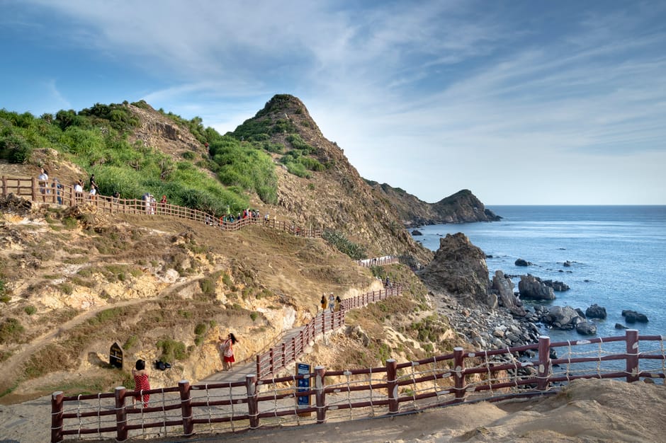 Travelers strolling on wooden pathway on rough grassy cliff slope washed by azure rippling sea on hot summer day