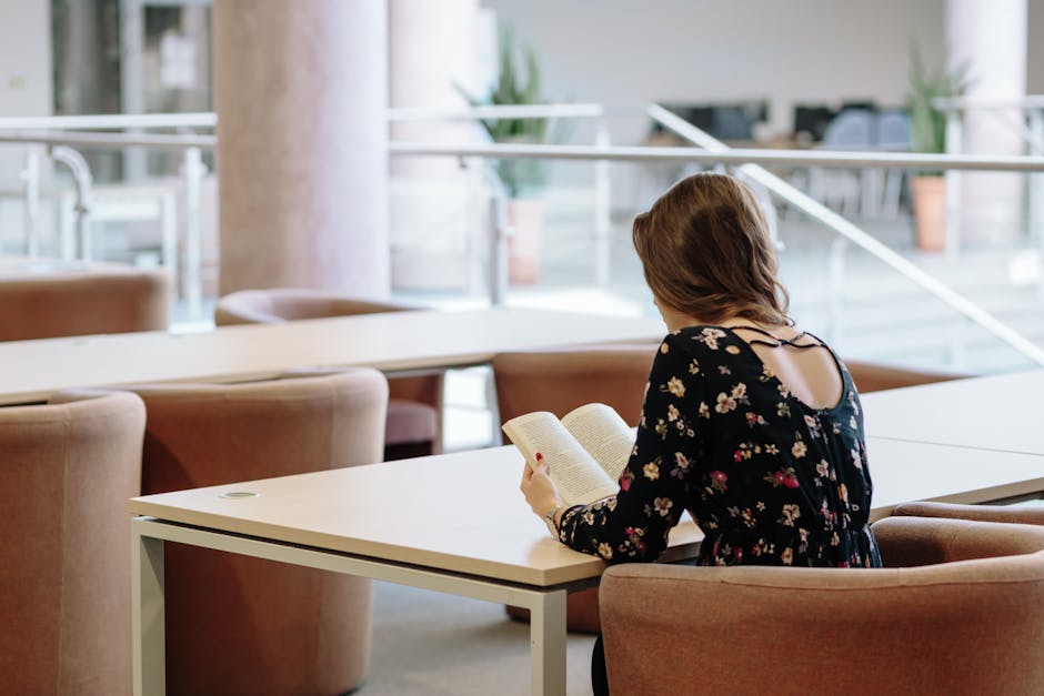 A woman sits comfortably indoors, reading a book in a modern library setting.