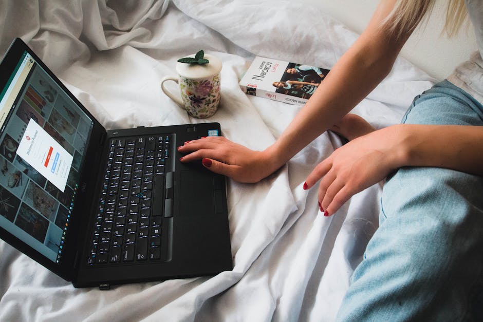 A person uses a laptop while resting on a bed, depicting a cozy work-from-home setup.