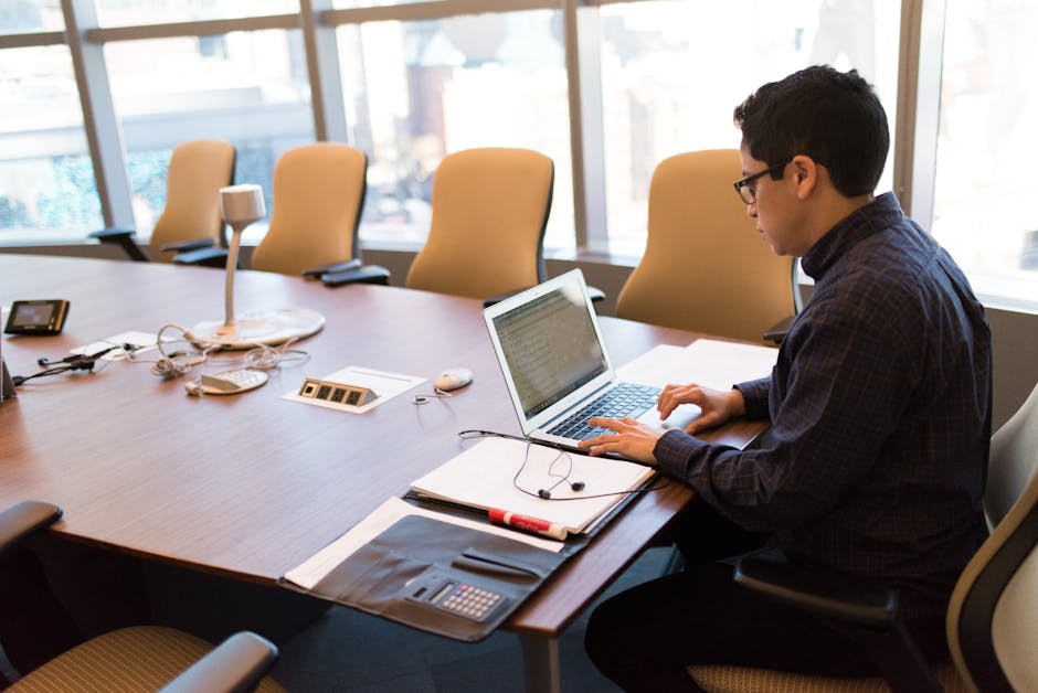 A professional adult sitting at a desk with a laptop in a bright, modern conference room.