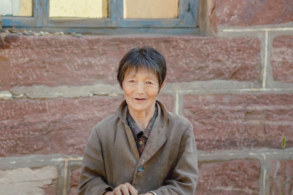 Charming portrait of an elderly Asian woman smiling against a rustic stone wall.