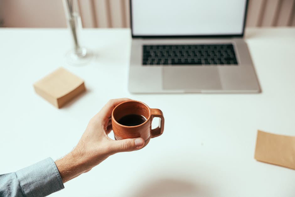 A person holds a brown coffee cup near a laptop on a clean white desk, perfect for remote work themes.