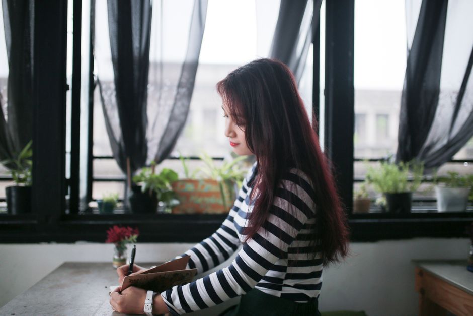 Side profile of a young woman writing in a stylish urban cafe with plants.