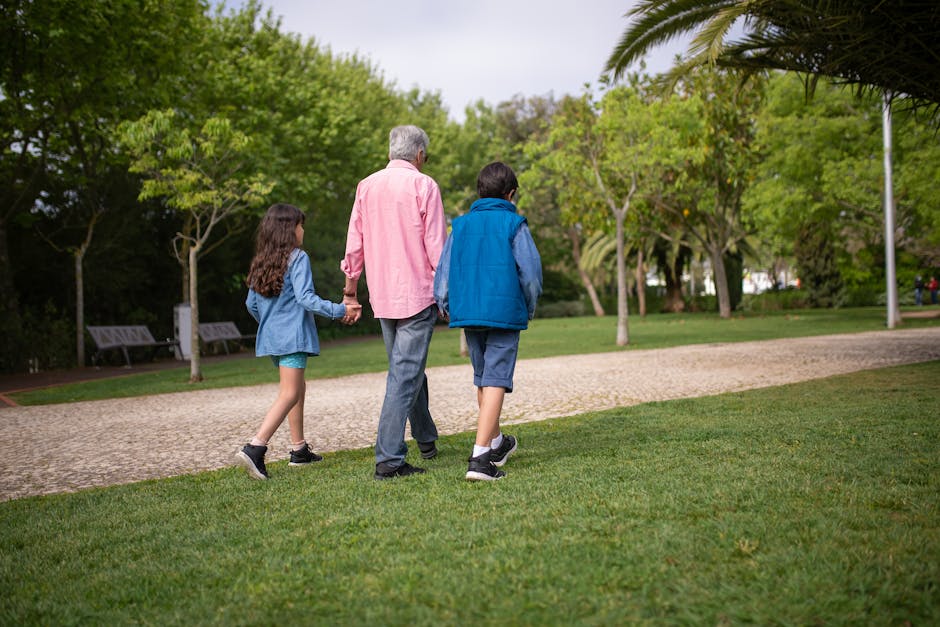 A grandfather holds hands with his grandchildren on a relaxing walk in a lush green park.