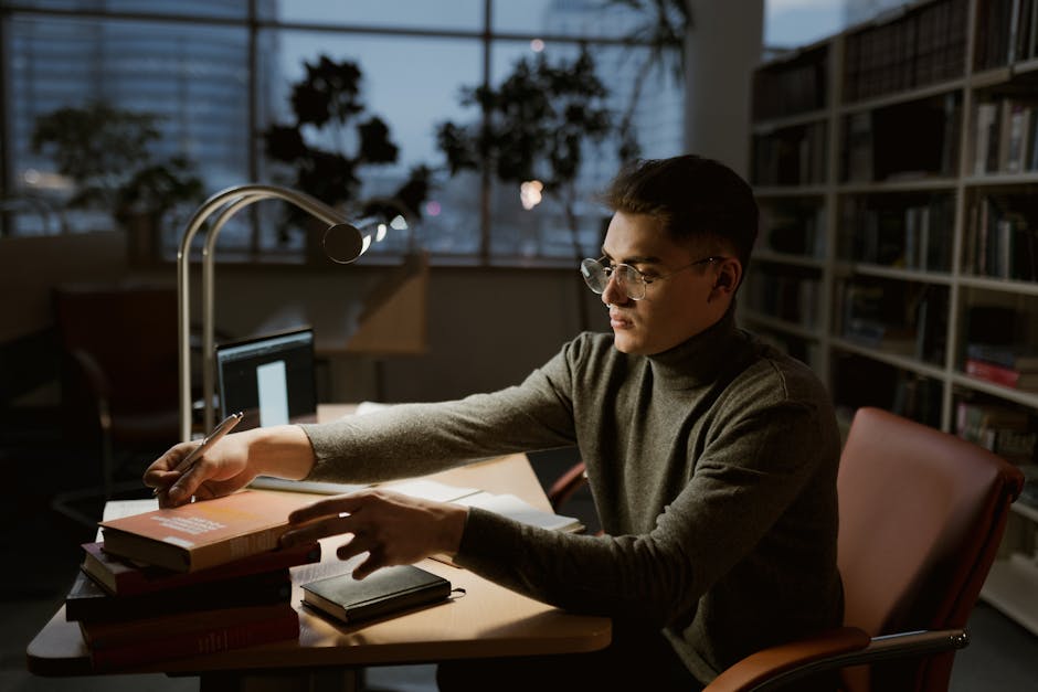 A young man wearing a turtleneck studies at a desk in a dimly lit library.