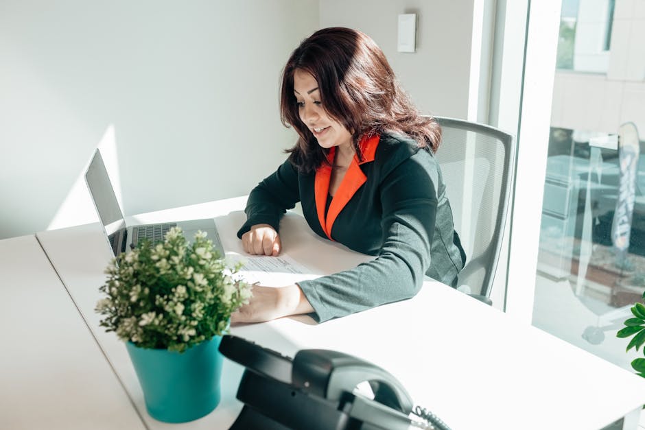 A woman in a bright office working at a desk with a laptop and plant.