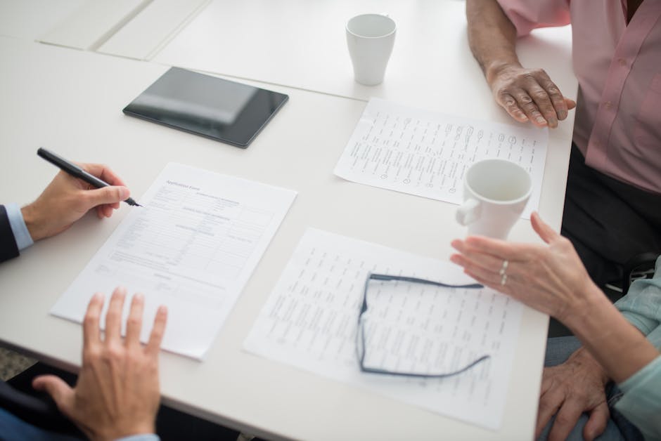 Hands writing on documents in a business meeting setup with coffee cups and tablet.