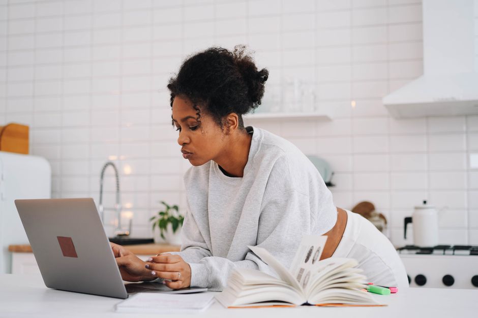 Woman at home in kitchen using laptop for online education with books around.