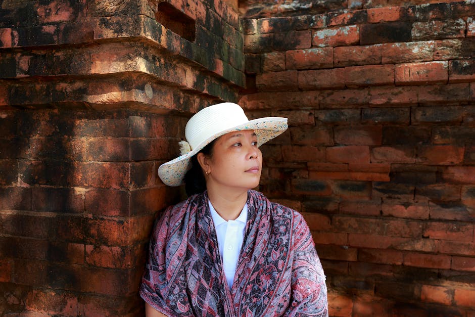 Woman wearing a white hat and scarf leans against a historical brick wall, gazing into the distance.