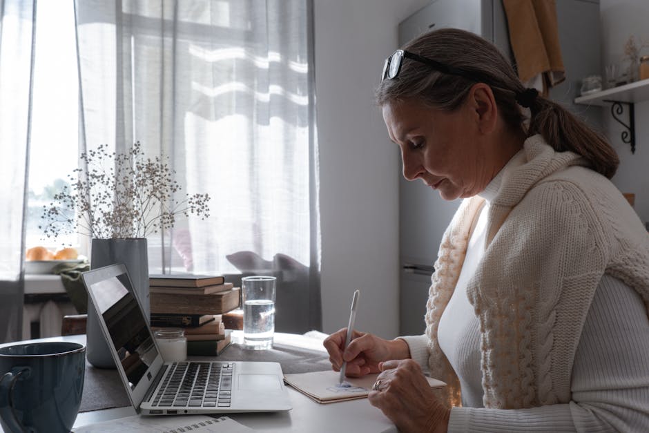 A woman in a knitted sweater writing notes at an indoor desk with a laptop.