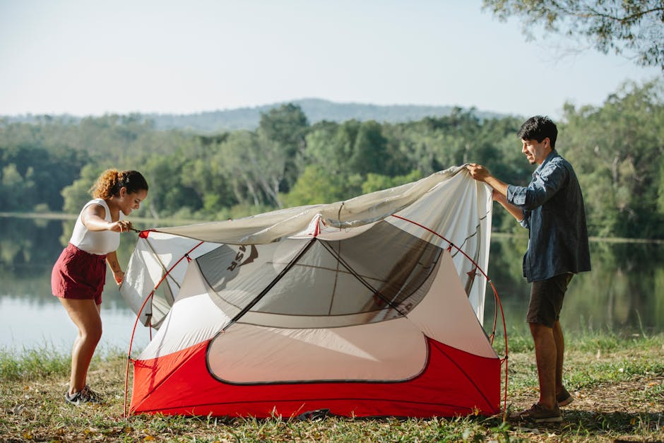 Full body side view of young multiracial couple of travelers putting up colorful tent on river bank covered with grass in summer day