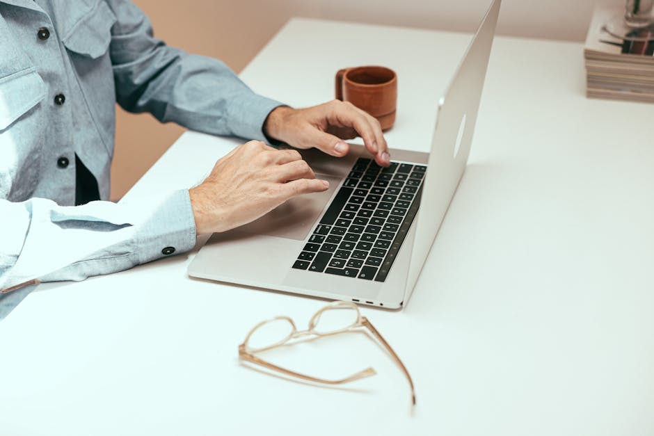 Adult working remotely on laptop with eyeglasses and cup on a white table.