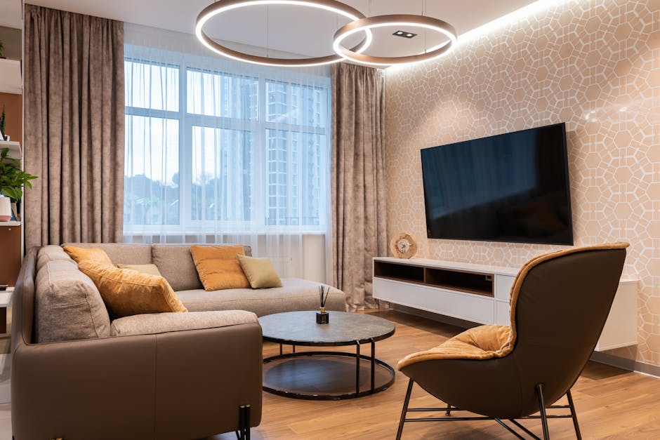 Interior of room with soft couch and pillows near window with curtains and armchair near round table and TV on wall near cabinet