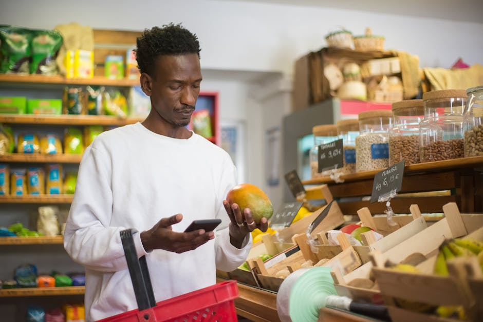 A man examines a mango while shopping with a smartphone in a grocery store.