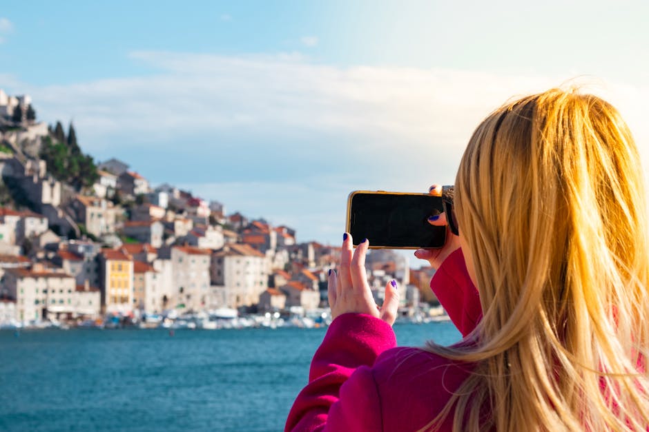 A woman takes a photo of the Šibenik waterfront using a smartphone on a sunny day.