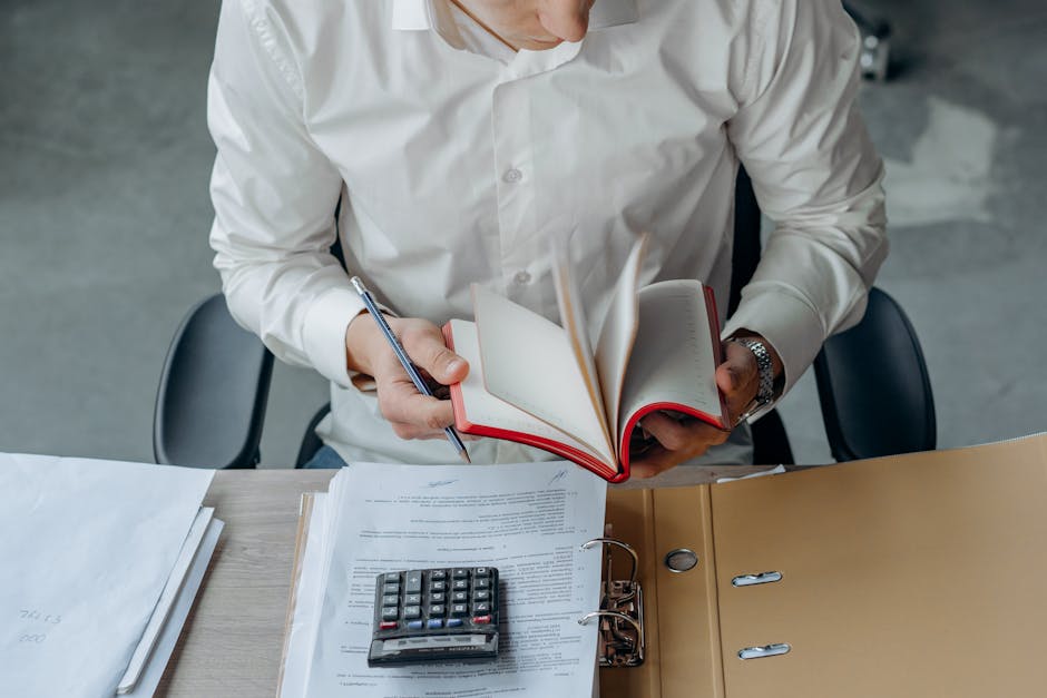 Man in white shirt reading book at office desk with calculator and documents.