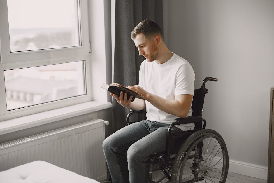 A man in a wheelchair reading a book in a well-lit room near a window.