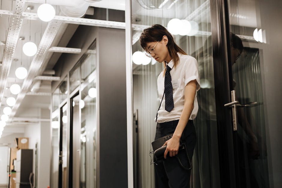 Young Asian adult leaning against a glass door in an office, appearing stressed, holding a tablet.