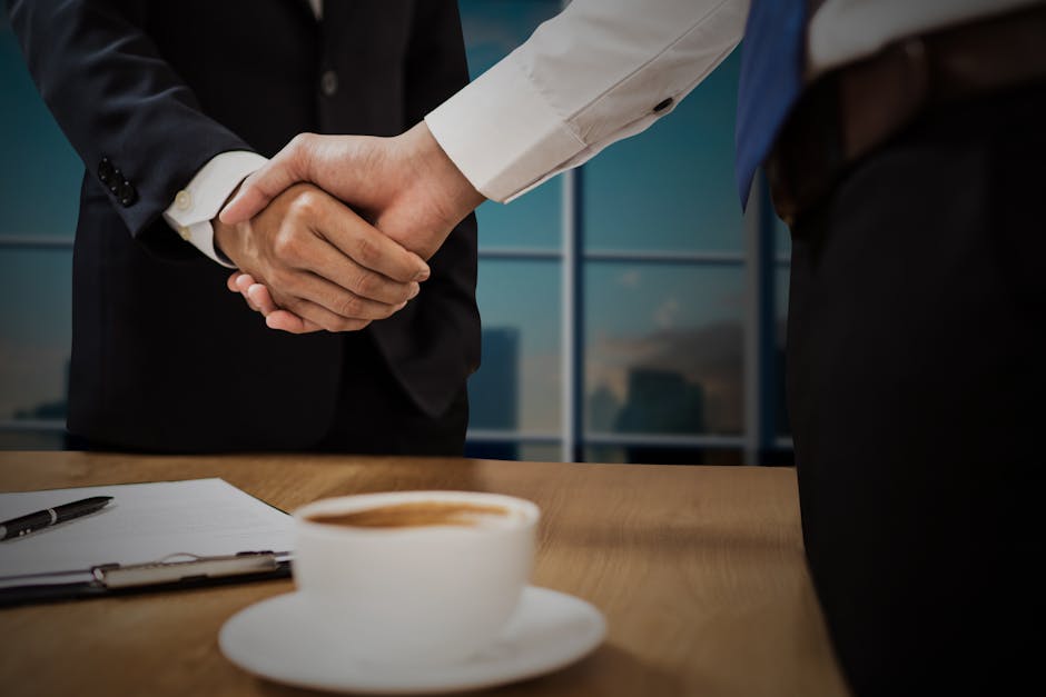 Two professionals shaking hands across a desk with cityscape in the background.