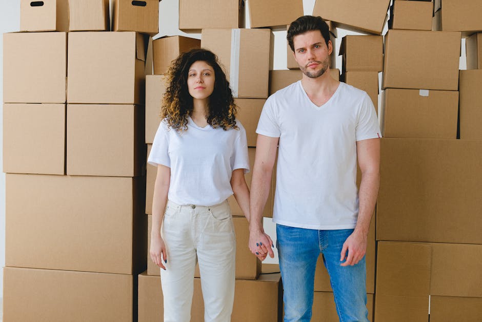 A young couple holding hands in front of a stack of cardboard boxes, possibly indicating moving or relocation.