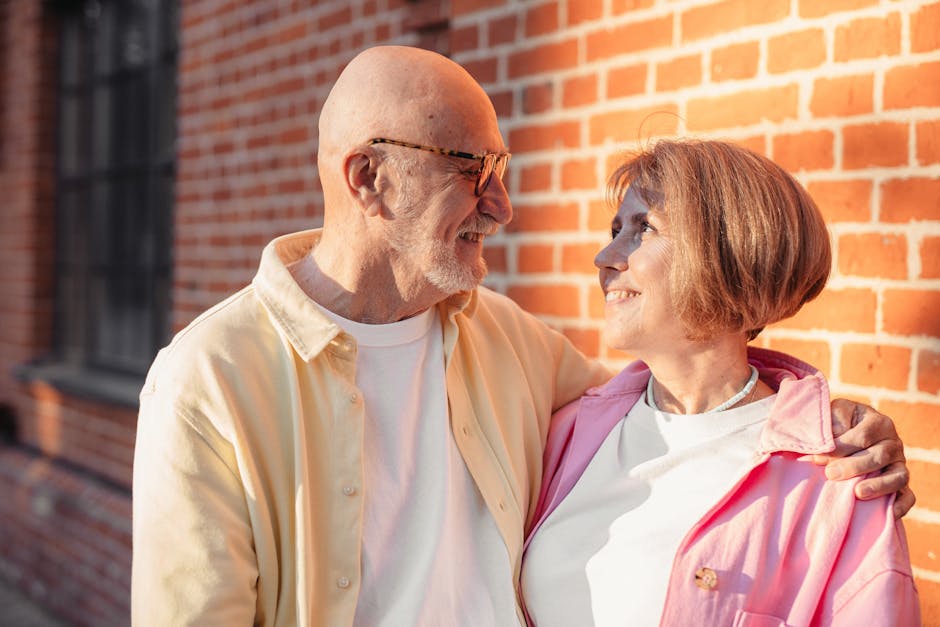 Elderly couple smiling and hugging against a sunlit brick wall, enjoying a moment of love.