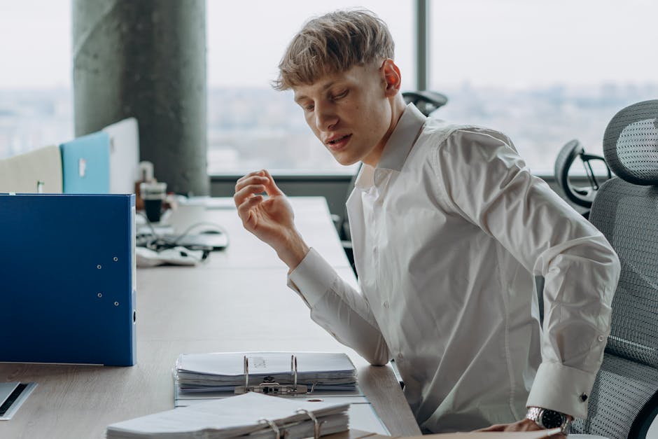 Man in white shirt sitting at office desk reading documents, modern workspace.