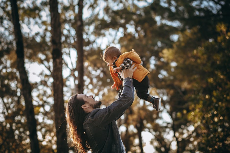 A father joyfully lifts his baby boy in a sunlit autumn park, surrounded by trees.