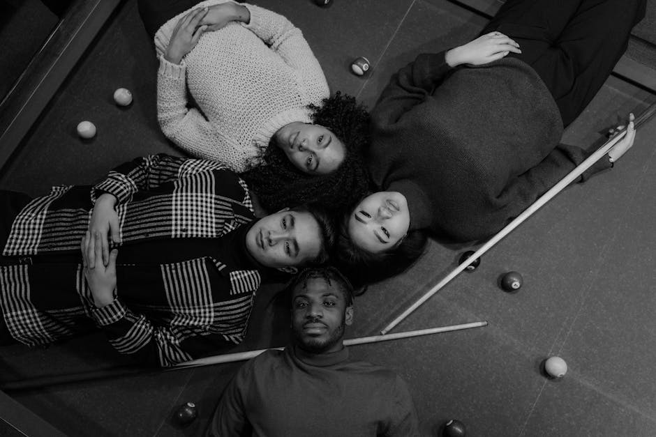 Four friends lie on a billiard table in a black-and-white photograph, sharing a moment of connection.