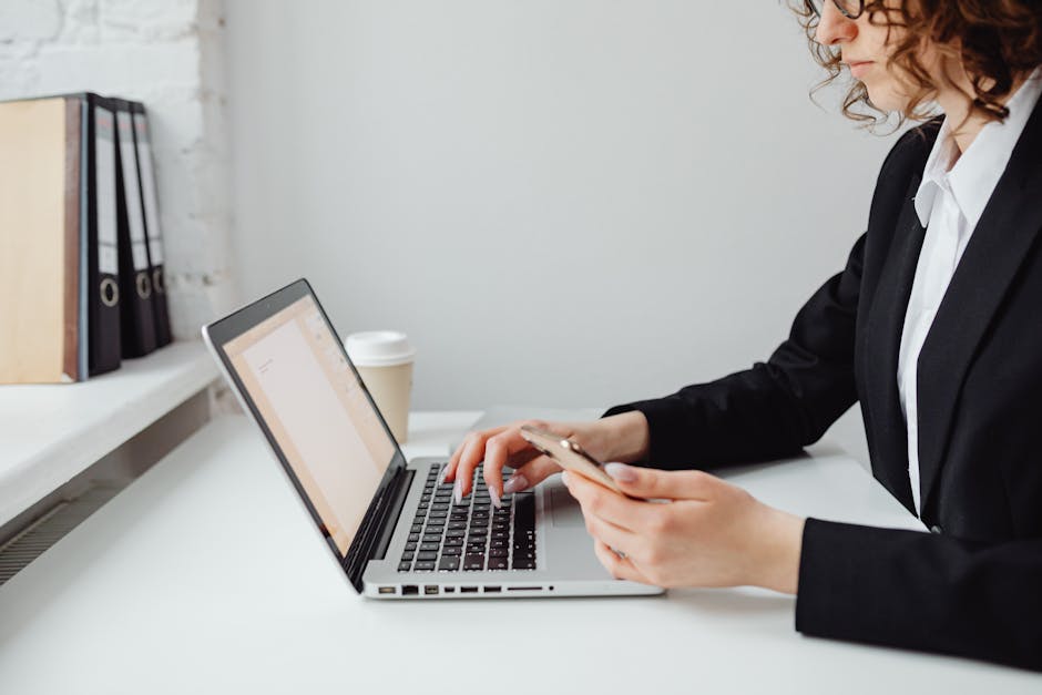 Professional woman in business attire using laptop and smartphone at a desk in an office setting.