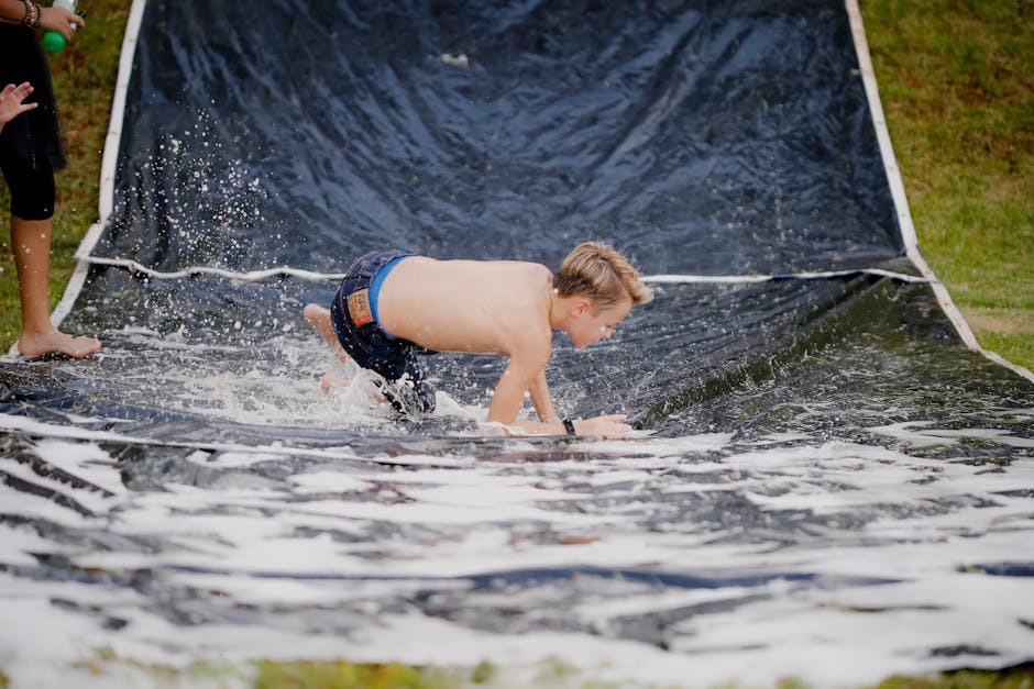 A young boy having fun on a slip and slide outside during a sunny day, splashing through water.