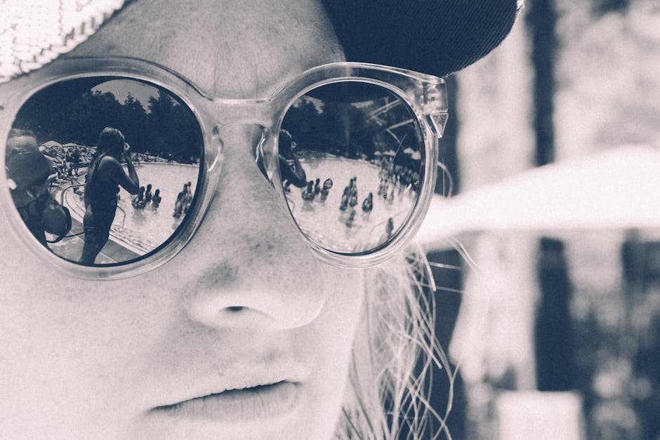 A woman wearing sunglasses reflects a busy poolside scene in black and white.