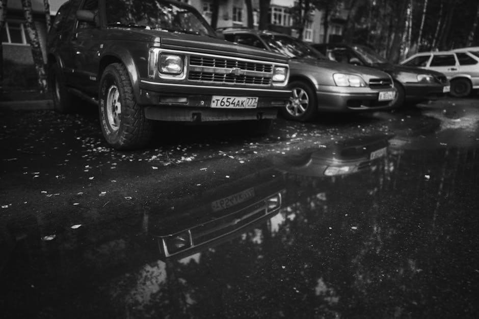 A dramatic black and white image of parked cars reflected in a rain puddle on a street.